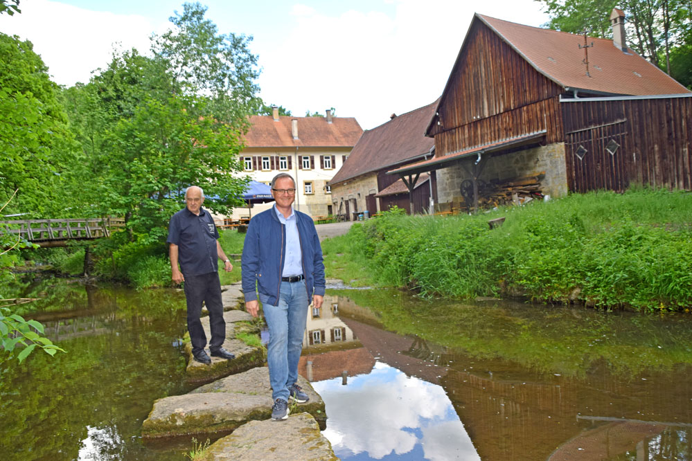 Reinhold Niebel (rechts) und Karl-Heinz Kurz auf den Steinen, die über die Gronach führen. Im Hintergrund ist die Hammerschmiede zu sehen. Die Steine liegen nicht zum Spaß im Fluss, sondern sind Markierungen für die daran angrenzende Furt. Bei genauem Blick ins Wasser kann man die Steinplatten am Grund erkennen. Foto: am